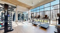 This fitness center is a thoughtfully designed, single-level facility offering a generous open workout space filled with abundant natural light. The most striking architectural feature is the expansive floor-to-ceiling glass curtain wall, framed in black steel, which spans the length of the room and provides unobstructed outdoor views while flooding the interior with daylight. Soaring ceilings are accented by warm wood-slat paneling and complemented by ceiling fans and recessed lighting, creating a bright and modern aesthetic.

The light-toned hardwood-style flooring runs throughout, contributing to a clean and contemporary look. The cardio zone is well-appointed with multiple treadmills, stationary bikes including Peloton cycles, ellipticals, and stair steppers, many positioned directly in front of the glass wall to enhance the workout experience with natural views. A dedicated strength training area features a multi-functional cable and power rack system, a comprehensive dumbbell and kettlebell rack, and a weight bench with free weights.

Large mirrors are positioned along interior walls to visually expand the space and support functional training. Black-framed interior glass partitions and doors connect the fitness area to adjacent spaces, contributing to the facility's cohesive, upscale design. The overall ambiance is energetic yet refined.