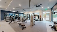 Located at 6302 Greenbelt Pkwy in Garland, TX, this fitness center offers a thoughtfully designed, single-floor exercise space that provides a comprehensive environment for a wide range of workout routines. The room features neutral-toned carpet flooring and crisp white walls accented by a bold navy blue feature wall, creating a modern and motivating atmosphere. Multiple ceiling fans are mounted throughout, ensuring comfortable air circulation during workouts. Recessed lighting illuminates the space evenly, complemented by abundant natural light streaming through large floor-to-ceiling windows that offer views of the surrounding greenery and streetscape. A full wall of mirrors expands the sense of space and allows users to monitor their form during exercise. The cardio area is equipped with a row of True treadmills and a stair climber, each featuring individual digital displays, alongside a wall-mounted television. The strength training section houses a variety of Hoist cable and selectorized machines, a multi-function cable trainer, a flat weight bench, and a well-stocked dumbbell rack. A dedicated area features a medicine ball rack with assorted weighted balls for functional training. Built-in shelving near the cardio section provides convenient storage for personal items, completing this fully outfitted community fitness facility.