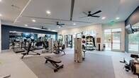 Located at 6302 Greenbelt Pkwy in Garland, TX, this fitness center offers a thoughtfully designed, single-floor exercise space that provides a comprehensive environment for a wide range of workout routines. The room features neutral-toned carpet flooring and crisp white walls accented by a bold navy blue feature wall, creating a modern and motivating atmosphere. Multiple ceiling fans are mounted throughout, ensuring comfortable air circulation during workouts. Recessed lighting illuminates the space evenly, complemented by abundant natural light streaming through large floor-to-ceiling windows that offer views of the surrounding greenery and streetscape. A full wall of mirrors expands the sense of space and allows users to monitor their form during exercise. The cardio area is equipped with a row of True treadmills and a stair climber, each featuring individual digital displays, alongside a wall-mounted television. The strength training section houses a variety of Hoist cable and selectorized machines, a multi-function cable trainer, a flat weight bench, and a well-stocked dumbbell rack. A dedicated area features a medicine ball rack with assorted weighted balls for functional training. Built-in shelving near the cardio section provides convenient storage for personal items, completing this fully outfitted community fitness facility.