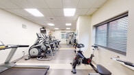 This fitness center is thoughtfully designed to support a wide range of exercise routines within a single-floor layout. The space features a drop-panel ceiling with recessed panel lighting that ensures consistent, bright illumination throughout. Neutral-toned walls are accented by a vibrant green feature wall, adding energy to the environment. A yellow floor stripe delineates cardio zones from open workout areas, contributing to an organized flow.

The cardio section is well-stocked with multiple Matrix treadmills, elliptical cross-trainers, and upright stationary bikes, including a dedicated spin cycle. Wall-mounted flat-screen televisions are positioned throughout for entertainment during workouts. A separate strength training area houses a multi-functional cable machine with pull-up bar attachments, an adjustable weight bench, and a rack of dumbbells, supported by instructional exercise posters on the walls.

Additional functional fitness equipment includes a floor-to-ceiling medicine ball rack in assorted weights, resistance bands, a foam roller, and a yoga mat, all neatly stored on wall-mounted hooks. A window with blinds provides natural light in the strength area. The space also features a wall-mounted air conditioning unit, ensuring a comfortable training environment year-round.