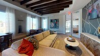 This thoughtfully designed single-floor amenity lounge offers a refined blend of comfort and entertainment. The space is anchored by a striking ceiling adorned with bold, dark-stained exposed wood beams that provide a warm architectural contrast against the neutral toned walls and soft carpeted flooring. Abundant natural light streams in through large floor-to-ceiling windows dressed with sheer white curtains, offering picturesque views of mature trees and a serene waterfront beyond.

The main lounge area features a generously sized sectional sofa accented with colorful throw pillows, complemented by sleek accent tables and contemporary artwork that add personality and visual interest throughout. A dedicated dining or game table surrounded by mid-century modern chairs serves as a central gathering point, ideal for casual social interaction.

A separate gaming zone includes a full-sized billiards table and a dedicated poker table surrounded by plush upholstered chairs, set beneath a coffered ceiling detail with built-in shelving niches for decorative storage. Glass partitions and doors connect adjoining spaces, enhancing the open, social atmosphere. The overall ambiance is sophisticated yet inviting, making this space well-suited for relaxed gatherings and recreational activities alike.