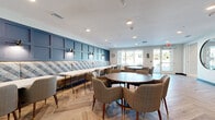 Located at 4351 Little Egret Rd in Kissimmee, Florida, this single-floor community clubhouse at Heron Ridge offers a well-appointed gathering space designed with both function and aesthetic appeal in mind. The entry foyer sets an inviting tone with herringbone-patterned wood-look tile flooring, a large round decorative mirror, and glass doors that flood the space with natural light and views of the outdoors. Throughout the space, the herringbone flooring creates a cohesive and stylish flow. The fully equipped kitchen features rich, dark wood cabinetry extending from floor to ceiling, complemented by clean white countertops and stainless steel appliances, including a full-size refrigerator and dishwasher. The open-plan lounge and dining area serves as the heart of the space, anchored by a striking blue paneled accent wall with built-in sconce lighting and an extended upholstered banquette in a plaid pattern. Multiple round dining tables with mid-century modern upholstered chairs provide comfortable and flexible seating arrangements. Large windows along the perimeter allow generous natural light to fill the room while offering pleasant outdoor views. Recessed lighting throughout ensures a well-lit, welcoming atmosphere. Nautical-themed artwork adds character and reinforces the community's coastal-inspired identity.