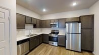 This single-floor space showcases a well-appointed kitchen that serves as a central highlight. The kitchen features a cohesive design with dark espresso-toned shaker-style cabinetry extending from floor to ceiling, providing generous storage through both upper and lower cabinets. The cabinetry is complemented by clean white countertops that create a striking contrast, lending a modern, polished aesthetic to the space.

The kitchen is fully equipped with a stainless steel appliance suite, including a gas range with oven, an over-the-range microwave, a full-size refrigerator, and a dishwasher. The layout follows an L-shaped configuration, offering a practical and efficient workflow. A stainless steel double-basin sink with a modern faucet is positioned along the perimeter, accompanied by a tasteful tile backsplash that adds a clean, finished look.

Light wood-style hard-surface flooring runs throughout, adding warmth and continuity to the space. Recessed lighting in the ceiling provides bright, even illumination throughout the kitchen. The white walls and neutral color palette contribute to an open, airy feel, making the space both functional and visually inviting.