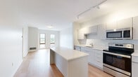 Located at 199 Rantoul St in Beverly, MA, this thoughtfully designed one-bedroom, one-bathroom unit offers a clean, contemporary aesthetic throughout its well-utilized floor plan. The open-concept kitchen and living area serves as the heart of the space, featuring sleek white cabinetry, a quartz-topped kitchen island, stainless steel appliances including a refrigerator, range, microwave, and dishwasher, and track lighting overhead. Light hardwood flooring runs seamlessly through the living and kitchen areas, enhancing the bright, modern feel. The living area is positioned adjacent to the kitchen, benefiting from natural light through dual windows that frame views of the surrounding Beverly neighborhood. The bedroom is a comfortable, well-proportioned room with large windows that invite natural light and offer outdoor views. It includes direct access to a dedicated walk-in closet fitted with wire shelving, providing practical storage. The full bathroom features large-format tile flooring, a floating vanity with a white countertop, and clean white walls, maintaining the minimalist design language found throughout the unit. With a neutral color palette, modern finishes, and an efficient layout, this unit offers a polished and functional living environment in the heart of Beverly, MA.