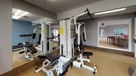 This single-floor fitness center is designed to provide a comprehensive workout environment with a practical, functional layout. The main exercise area is outfitted with a multi-station strength training machine, a treadmill, a stationary recumbent bike, and an upright cycle, offering a well-rounded selection of equipment for both cardio and resistance training. Neutral-toned carpet flooring runs throughout, providing a comfortable surface underfoot. Large floor-to-ceiling mirrors line one wall, enhancing the sense of space and allowing users to monitor their form during workouts. The walls are finished in a calm blue-grey palette, contributing to a focused yet relaxed atmosphere. Fluorescent overhead lighting ensures the space is consistently well-lit. Double French doors along one wall invite natural light into the room and provide direct exterior access, offering views of the surrounding outdoor area. A pass-through window connects the fitness room to an adjacent lounge space, which features light hardwood-style flooring, recessed lighting, and comfortable seating arrangements. This thoughtful connection between the fitness area and the lounge creates a cohesive, multi-purpose community facility suited for both active use and relaxed social interaction.
