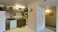 This single-floor space presents a practical and straightforward layout centered around a well-equipped kitchen. The kitchen features dark wood cabinetry providing ample upper and lower storage, complemented by a dark-toned countertop housing a double-basin sink. Appliances include a white gas range, over-the-range microwave, and a dishwasher, all finished in white for a cohesive look. Overhead fluorescent lighting illuminates the workspace effectively. A decorative rug adds a touch of warmth to the light-colored flooring. Adjacent to the kitchen, a hallway connects to additional areas of the home, highlighted by a set of white double doors with paneled detailing, likely concealing a closet or storage space. The walls throughout are painted in a neutral, light tone, creating a clean and airy atmosphere. A wall clock and overhead HVAC vent are visible, adding to the functional character of the space. The overall design prioritizes utility, making it a practical living environment suited for everyday use.