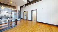 Unit 205 at 101 E Sixth Street is an urban loft-style residence in downtown Winston-Salem, NC, offering two bedrooms and two bathrooms across a single open floor. The space showcases industrial-chic design with warm hardwood floors throughout, soaring ceilings, and exposed ductwork. Large multi-pane factory-style windows flood the main living area with natural light and offer views of the surrounding cityscape. The kitchen features granite countertops, rich dark wood cabinetry, a peninsula with open shelving, and pendant lighting. Both bedrooms are well-sized and share the same warm hardwood flooring. The two bathrooms are finished with tile flooring, full tub-shower combinations, and clean modern fixtures, providing functional and comfortable amenities throughout.