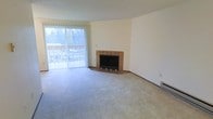 This single-floor space offers a comfortable and functional layout centered around a generously sized living area. The room is finished with soft wall-to-wall carpeting that adds warmth underfoot, while neutral white walls provide a clean, versatile backdrop suited to a variety of décor styles. A standout feature of the living space is the classic brick fireplace, positioned in the corner and framed by a tiled hearth, serving as a natural focal point that brings both character and coziness to the room. Opposite the fireplace, a large sliding glass door with vertical blinds allows an abundance of natural light to pour in, creating a bright and airy atmosphere throughout the day. The sliding door also provides direct access to an outdoor area, offering views of the surrounding greenery. A baseboard heating unit along the wall ensures year-round comfort. The overall ambiance of the space is calm and understated, with practical finishes that make it move-in ready and easy to personalize. The open feel of the main living area makes it well-suited for both everyday living and casual entertaining.