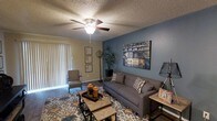 This single-story space centers around a well-appointed living room that combines comfort and style. A striking slate-blue accent wall serves as a focal point, adorned with a large canvas artwork that adds visual interest and depth to the room. The remaining walls are finished in a neutral beige tone, creating a balanced and inviting contrast.

The living room is furnished with a gray upholstered sofa dressed with patterned throw pillows, complemented by a matching accent chair positioned near the sliding glass door. A patterned area rug in earth tones and blue anchors the seating arrangement, while a set of wood-and-metal coffee tables adds a rustic, layered touch. A floor lamp and side table provide functional lighting and surface space.

Overhead, a ceiling fan with an integrated light fixture is mounted on a textured popcorn ceiling, offering both air circulation and ambient illumination. Vertical blinds cover the sliding glass door, which allows for natural light and provides access to an outdoor area.

The overall ambiance is casual yet thoughtfully styled, with a mix of textures and tones that create a warm and livable atmosphere throughout the space.