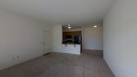 This single-floor space offers an open-concept design that seamlessly connects the living area and kitchen, creating a practical and cohesive layout. The living area is finished with wall-to-wall carpeting in a neutral tone, providing a comfortable underfoot feel while contributing to a clean, understated aesthetic throughout the space.

The kitchen is positioned centrally and defined by a white peninsula counter that doubles as a breakfast bar, offering casual seating and additional workspace. Warm-toned wooden cabinetry lines the upper portion of the kitchen, providing ample storage. The kitchen is equipped with black appliances, including a refrigerator and an over-the-range microwave, alongside a sink integrated into the peninsula. Recessed and flush-mount ceiling lights illuminate both the kitchen and the adjoining areas effectively.

A white paneled entry door is situated along one wall, offering a clear and accessible entry point into the living space. The walls throughout are painted in a crisp white, enhancing the brightness of the interior. The overall layout is straightforward and well-organized, making efficient use of the available space on a single level.