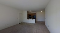 This single-floor space offers an open-concept design that seamlessly connects the living area and kitchen, creating a practical and cohesive layout. The living area is finished with wall-to-wall carpeting in a neutral tone, providing a comfortable underfoot feel while contributing to a clean, understated aesthetic throughout the space.

The kitchen is positioned centrally and defined by a white peninsula counter that doubles as a breakfast bar, offering casual seating and additional workspace. Warm-toned wooden cabinetry lines the upper portion of the kitchen, providing ample storage. The kitchen is equipped with black appliances, including a refrigerator and an over-the-range microwave, alongside a sink integrated into the peninsula. Recessed and flush-mount ceiling lights illuminate both the kitchen and the adjoining areas effectively.

A white paneled entry door is situated along one wall, offering a clear and accessible entry point into the living space. The walls throughout are painted in a crisp white, enhancing the brightness of the interior. The overall layout is straightforward and well-organized, making efficient use of the available space on a single level.