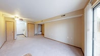 This single-floor space presents a practical and straightforward layout designed for comfortable everyday living. The main living area is generously sized, featuring light beige carpet flooring that extends across the room, contributing to a warm and understated aesthetic. Neutral-toned walls in a soft tan hue are complemented by white ceilings, creating a clean and cohesive look throughout.

The living area is well-ventilated, with multiple ceiling vents visible, ensuring consistent airflow throughout the space. Wood-toned baseboards and door trim provide a subtle contrast to the light walls, adding a modest architectural detail. Multiple electrical outlets are conveniently positioned along the walls, offering practical utility for furniture arrangement.

A doorway at the far end of the living area leads to additional rooms, suggesting a connected and functional floor plan. To the left, a partial view of what appears to be a kitchen area is visible, featuring light cabinetry. A solid wood-toned door nearby likely leads to a utility or storage area. The overall ambiance of the space is neutral, clean, and move-in ready, offering a blank canvas for personalization.