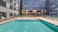 This contemporary residential building showcases a thoughtfully designed courtyard amenity space enclosed by a multi-story façade finished in contrasting gray horizontal siding and warm wood-toned panel cladding. Large, dark-framed windows line the building's exterior, offering abundant natural light to the units within while overlooking the vibrant courtyard below.

At the heart of the outdoor space is a generously sized swimming pool with clear blue water, complete with an accessibility lift and lined by a concrete deck outfitted with lounge chairs. Elegant white pergola structures provide shaded retreat areas around the pool, adding architectural definition to the space.

Adjacent to the pool area, a dedicated lawn zone features lush artificial turf beneath a wood-beamed pergola with string lights, creating an inviting and relaxed atmosphere. A hanging wicker egg chair adds a stylish focal point to this recreational area.

Thoughtfully curated landscaping is woven throughout the courtyard, with raised planting beds, ornamental shrubs, ground cover, and planters softening the hardscape and enhancing the overall ambiance. Paved walkways connect the various zones seamlessly. A covered entry vestibule with metal railings and planters provides a transitional passage into the building. The entire amenity level reflects a cohesive modern design aesthetic with attention to both functionality and resident comfort.