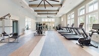 Located in Santa Rosa Beach, FL, this fitness center offers a generously sized, single-floor exercise space designed for a comprehensive workout experience. The room's architecture is immediately striking, with soaring ceilings adorned by rustic exposed wooden beams and a coffered ceiling detail that adds both warmth and visual depth. Large, floor-to-ceiling windows line the walls, flooding the interior with natural light and providing open views of the surrounding neighborhood.

The exercise floor is thoughtfully zoned, with durable flooring that transitions between smooth hardwood-style panels and multi-toned area rugs, clearly delineating cardio and strength training areas. A row of treadmills and stationary bikes is positioned along the windowed wall, allowing users to exercise with an outdoor view. The strength training zone features cable machines, a weight bench, a squat rack, stability balls, and a full rack of dumbbells, catering to a wide range of fitness routines.

Recessed lighting and a large pendant ceiling fan ensure the space remains well-lit and comfortable. French doors on one end provide additional access and airflow. The overall ambiance is bright, clean, and inviting, combining coastal-inspired design with practical, high-quality fitness amenities in a polished, community-oriented setting.