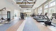 Located in Santa Rosa Beach, FL, this fitness center offers a generously sized, single-floor exercise space designed for a comprehensive workout experience. The room's architecture is immediately striking, with soaring ceilings adorned by rustic exposed wooden beams and a coffered ceiling detail that adds both warmth and visual depth. Large, floor-to-ceiling windows line the walls, flooding the interior with natural light and providing open views of the surrounding neighborhood.

The exercise floor is thoughtfully zoned, with durable flooring that transitions between smooth hardwood-style panels and multi-toned area rugs, clearly delineating cardio and strength training areas. A row of treadmills and stationary bikes is positioned along the windowed wall, allowing users to exercise with an outdoor view. The strength training zone features cable machines, a weight bench, a squat rack, stability balls, and a full rack of dumbbells, catering to a wide range of fitness routines.

Recessed lighting and a large pendant ceiling fan ensure the space remains well-lit and comfortable. French doors on one end provide additional access and airflow. The overall ambiance is bright, clean, and inviting, combining coastal-inspired design with practical, high-quality fitness amenities in a polished, community-oriented setting.
