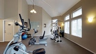 This fitness center offers a comfortable and functional environment for residents to maintain an active lifestyle. The space is defined by its striking vaulted ceiling, which creates an open, airy atmosphere and adds a sense of spaciousness. A classic chandelier hangs centrally, complementing the natural light that streams in through multiple large windows fitted with blinds along the far wall, keeping the space bright and inviting.

The neutral-toned walls and carpeted flooring contribute to a clean, welcoming aesthetic throughout. The layout is thoughtfully arranged to accommodate a variety of workout needs. Cardio equipment, including upright and recumbent stationary bikes, is positioned toward the front of the room, while a multi-function cable strength training machine occupies the center of the space. An exercise ball and additional equipment are visible along the perimeter, providing options for flexibility and supplemental training. Framed artwork on the walls and a small seating area add a refined touch to the functional setting. The overall ambiance balances practicality with a warm, residential feel, making it an accessible and pleasant space for daily exercise.