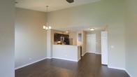 This single-floor residence offers a well-designed open-concept layout that maximizes both space and functionality. The living and dining areas flow naturally into one another, anchored by rich, dark hardwood flooring that extends throughout the main living space. Soaring ceilings lend an airy, expansive feel to the interior, enhanced by a stylish chandelier that serves as a focal point above the dining area.

The kitchen is positioned at the heart of the home, featuring dark cabinetry, stainless steel appliances including a microwave and refrigerator, and a light-toned countertop peninsula that doubles as a breakfast bar. This peninsula creates a natural visual separation between the kitchen and the living area while maintaining the open feel of the floor plan.

The neutral, warm-toned walls complement the contrast between the dark cabinetry and flooring, creating a cohesive and modern aesthetic throughout. A ceiling fan overhead adds practical comfort to the space. A paneled entry door visible from the main living area provides convenient access, further highlighting the thoughtful and accessible layout of this single-level home. Overall, the space strikes a balance between contemporary design and everyday livability.
