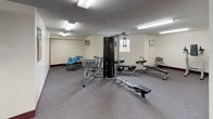 This fitness center offers a comprehensive workout environment across a single open floor. The space is generously sized, providing ample room to move freely between equipment stations without feeling crowded. Flooring throughout is covered in a durable, speckled carpet, while the walls are finished in a clean off-white tone with a deep burgundy baseboard accent that runs the perimeter of the room.

The cardio area is well-appointed, featuring multiple treadmills, elliptical trainers, and a stationary recumbent bike. A large wall-to-wall mirror spans one side of the room, visually expanding the space and allowing users to monitor their form during workouts. The strength training zone includes a multi-station cable machine, a rowing bench, an inversion table, and additional resistance equipment.

Overhead, rows of fluorescent panel lighting provide consistent, even illumination throughout. A wall-mounted television is positioned for easy viewing from the cardio area. A vending machine is conveniently located near the entrance. The textured ceiling adds a classic finish to the room. A standard access door provides entry, and a wall-mounted air conditioning unit helps maintain a comfortable environment. Overall, the fitness center is practical, functional, and equipped to support a variety of workout routines.