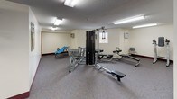 This fitness center offers a comprehensive workout environment across a single open floor. The space is generously sized, providing ample room to move freely between equipment stations without feeling crowded. Flooring throughout is covered in a durable, speckled carpet, while the walls are finished in a clean off-white tone with a deep burgundy baseboard accent that runs the perimeter of the room.

The cardio area is well-appointed, featuring multiple treadmills, elliptical trainers, and a stationary recumbent bike. A large wall-to-wall mirror spans one side of the room, visually expanding the space and allowing users to monitor their form during workouts. The strength training zone includes a multi-station cable machine, a rowing bench, an inversion table, and additional resistance equipment.

Overhead, rows of fluorescent panel lighting provide consistent, even illumination throughout. A wall-mounted television is positioned for easy viewing from the cardio area. A vending machine is conveniently located near the entrance. The textured ceiling adds a classic finish to the room. A standard access door provides entry, and a wall-mounted air conditioning unit helps maintain a comfortable environment. Overall, the fitness center is practical, functional, and equipped to support a variety of workout routines.