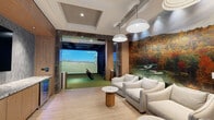 This indoor golf simulator suite offers a refined, amenity-rich environment designed for both practice and entertainment. The space is divided into two distinct zones: a dedicated simulator bay and an elegantly appointed lounge area, seamlessly connected through a dramatic dark marble-clad archway.

The simulator bay is fully equipped with a large high-definition projection screen, turf flooring with a designated hitting mat, and a ceiling-mounted projector that delivers immersive, course-realistic gameplay. A built-in control station with a touchscreen display and storage cabinetry provides easy access to simulator settings.

The adjacent lounge area is thoughtfully designed for relaxation and socializing. Plush neutral-toned armchairs are arranged around round pedestal side tables, all set against a striking floor-to-ceiling photographic mural depicting an autumn golf course landscape. Pendant lighting with layered white shades casts a warm, inviting glow, while light hardwood flooring and coffered ceiling details elevate the overall aesthetic.

A sleek wet bar runs along one wall, featuring granite countertops, wood cabinetry, an undermount sink, a wall-mounted television, and a built-in beverage cooler. A frosted glass entry door with a black frame completes the polished, contemporary design, making this suite an exceptional space for golf enthusiasts and entertainers alike.