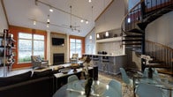 This thoughtfully designed single-floor space makes a strong impression with its dramatically high ceilings, which lend an expansive, airy quality to the interior. The open-concept layout connects the main living area, dining space, and a built-in bar counter seamlessly, encouraging a natural flow throughout. The living area is anchored by a striking stone-clad fireplace flanked by tall windows with vivid orange-trimmed frames, flooding the space with natural light while adding a bold architectural accent. Track lighting and pendant fixtures illuminate the space from above, complementing the ceiling fan centered overhead. The bar area features a raised countertop with barstool seating, mosaic tile backsplash detailing, and a small beverage station, making it a functional and stylish focal point. A glass-top dining table with modern chairs occupies the foreground, adding a contemporary touch. A wrought-iron spiral staircase rises near the bar area, adding vertical interest and an industrial-chic element. Upper shelving near the staircase provides decorative display space. The overall ambiance is modern yet inviting, blending warm textures with clean lines.