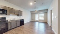 This single-floor unit showcases a thoughtfully designed open-concept layout that seamlessly connects the kitchen and living areas. The kitchen is well-appointed with dark taupe cabinetry featuring brushed nickel hardware, light countertops, and a full suite of black appliances including a refrigerator, range, microwave, dishwasher, and sink. Wood-style hard surface flooring runs throughout the main living areas, providing a warm and cohesive aesthetic.

The living area is generously proportioned, benefiting from natural light that streams through a set of double doors fitted with white blinds. A ceiling fan is centrally positioned, providing both comfort and additional lighting. The neutral wall tones throughout create a clean, modern atmosphere.

The bedroom offers plush carpeting, a ceiling fan, and a window with blinds that admits natural light, contributing to a comfortable and restful environment.

The bathroom is well-sized and features a wide vanity with dark wood cabinetry, ample drawer and cabinet storage, a large mirror with overhead lighting, a single-basin sink with light countertops, and wood-style flooring that mirrors the rest of the unit. The overall design is consistent and contemporary throughout.