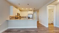 This single-floor residence at 1450 Upper Manatee River Rd in Bradenton, Florida offers a clean, contemporary design throughout. The open-concept kitchen serves as the centerpiece of the living area, featuring white shaker-style cabinetry extending to the ceiling, a tiled backsplash with under-cabinet lighting, and granite countertops. A spacious peninsula with pendant lighting above provides additional workspace and casual seating. Stainless steel appliances, including a French door refrigerator with water and ice dispenser, electric range, and over-the-range microwave, complete the well-appointed kitchen. Wood-look luxury vinyl plank flooring runs consistently throughout the space, lending warmth and visual continuity. A bright dining area adjacent to the kitchen benefits from natural light through a window with white blinds. The hallway connects the living spaces to the private areas, where a bathroom features a granite-topped white vanity, a large mirror, and access to a walk-in closet with built-in shelving. A dedicated laundry area accommodates a full-size front-loading washer and includes overhead wire shelving for storage. The overall palette of soft neutrals, white trim, and recessed lighting creates a bright, cohesive ambiance throughout.