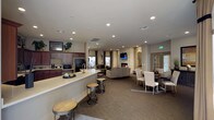 This single-floor space is designed as a versatile, open-concept gathering area that seamlessly integrates multiple functional zones. The kitchen area features dark wood cabinetry, a black refrigerator, built-in microwave, and a spacious white countertop bar with industrial-style round stools, providing a casual seating option for socializing or light refreshments. Recessed lighting throughout the ceiling ensures consistent, well-distributed illumination across the entire space.

The central lounge zone is anchored by a striking stacked-stone fireplace with a wall-mounted television above it, creating a warm and inviting focal point. Comfortable neutral-toned seating is arranged to encourage conversation and relaxation around the fireplace. Adjacent to the lounge, a formal dining area features upholstered white chairs around a dark wood table, offering a more structured setting for meals or meetings. Natural light enters through windows near the dining and lounge areas, complementing the recessed lighting. Neutral carpet flooring and warm wall tones contribute to a cohesive, welcoming ambiance throughout the space.