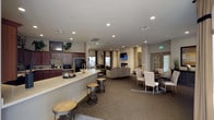 This single-floor space is designed as a versatile, open-concept gathering area that seamlessly integrates multiple functional zones. The kitchen area features dark wood cabinetry, a black refrigerator, built-in microwave, and a spacious white countertop bar with industrial-style round stools, providing a casual seating option for socializing or light refreshments. Recessed lighting throughout the ceiling ensures consistent, well-distributed illumination across the entire space.

The central lounge zone is anchored by a striking stacked-stone fireplace with a wall-mounted television above it, creating a warm and inviting focal point. Comfortable neutral-toned seating is arranged to encourage conversation and relaxation around the fireplace. Adjacent to the lounge, a formal dining area features upholstered white chairs around a dark wood table, offering a more structured setting for meals or meetings. Natural light enters through windows near the dining and lounge areas, complementing the recessed lighting. Neutral carpet flooring and warm wall tones contribute to a cohesive, welcoming ambiance throughout the space.