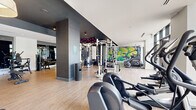 Located at 780 S 52nd St in Philadelphia's 19143 neighborhood, this contemporary fitness center offers a generously proportioned exercise space designed with both function and aesthetics in mind. The single-floor layout is finished with light wood-look flooring throughout, providing a clean and durable surface suited to high-traffic workouts. Dark charcoal accent walls and bold black ceiling details contrast with bright white walls and recessed lighting, creating a striking, modern atmosphere. A standout artistic mural featuring vibrant graffiti-style artwork serves as a dynamic focal point, adding an urban energy to the space. Floor-to-ceiling glass windows and sliding doors flood the cardio area with abundant natural light and offer open views to an adjoining outdoor terrace, seamlessly blending interior and exterior environments. The fitness floor is comprehensively equipped, featuring a cable and pulley multi-station, a Smith machine with bench press, a full dumbbell rack, plyo boxes, a rowing machine, elliptical trainers, stationary cycling bikes with integrated displays, and multiple treadmills. Large mirrors are strategically placed to expand the sense of space and allow users to monitor their form. Overhead pendant lighting adds a refined touch, while the overall layout ensures ample open floor space for functional and free-form training.