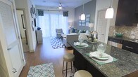 This single-floor space showcases an open-concept layout that seamlessly connects the kitchen and living areas, creating a cohesive and functional environment. The kitchen features a prominent breakfast bar with rich, dark granite countertops and pendant lighting overhead, offering casual seating on bar-height stools — an ideal setup for dining or entertaining. The countertop's deep tones contrast elegantly with the lighter surroundings.

Hardwood-style flooring extends throughout the space, lending warmth and a sense of continuity. The living area is generously proportioned, accommodating a full seating arrangement centered around a bold geometric area rug in blue and white tones. A ceiling fan with integrated lighting enhances comfort and airflow.

Large sliding glass doors with vertical blinds and decorative teal drapery anchor the far wall, flooding the interior with natural light and providing access to an outdoor area. The color palette throughout combines cool teal and grey accents with neutral furniture, contributing to a modern and inviting ambiance. Wall décor and layered lighting add personalized character to the overall design.