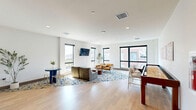 Located at 1201 Callowhill St in Philadelphia's Callowhill neighborhood, this contemporary amenity lounge offers a well-appointed, open-plan layout finished with light hardwood flooring throughout. The living area is anchored by large, black-framed windows that frame sweeping views of the surrounding Philadelphia cityscape, flooding the space with abundant natural light. Comfortable seating arrangements, including a leather lounge chair and sofa, are set atop a bold blue, gold, and grey patterned area rug, creating a warm and inviting atmosphere. A compact dining table with chairs provides an intimate spot for casual gatherings, while a wall-mounted television adds a modern entertainment element. The social side of the space features a full-length shuffleboard table flanked by bar-height chairs, set beneath a curated gallery of colorful abstract artwork — ideal for recreation and community engagement. The staircase area stands out as an architectural highlight, showcasing sleek black metal railings with horizontal cable detailing, warm wood treads, and a skylight above that draws in additional natural light. A built-in counter niche with blue tile accents adds a refined, functional touch near the stair landing. Clean white walls and recessed lighting complete the polished, modern aesthetic throughout.