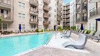 Located at 4015 Craft St in Charlotte, NC, this outdoor amenity courtyard is set within a modern multi-story residential complex and offers a well-appointed communal retreat. The centerpiece of the space is a generously sized resort-style swimming pool with crystal-clear blue water, surrounded by a broad paver pool deck furnished with vibrant red and neutral lounge chairs, inviting relaxation throughout the day. A dedicated shallow sun shelf with contemporary molded chaise lounges adds an additional layer of leisure to the poolside experience. Adjacent to the pool, a landscaped garden corridor winds through the courtyard, featuring raised brick planter beds filled with lush ornamental grasses, flowering plants, and mature shrubs, all framed by a paved walkway lined with modern outdoor furniture. A built-in wood-topped concrete fire or prep table serves as a functional outdoor gathering feature, complemented by sleek seating. At one end of the courtyard, a sophisticated clubhouse entrance is defined by floor-to-ceiling steel-framed glass windows and brick façade detailing, with potted greenery and casual Adirondack-style seating creating a welcoming transition between the interior amenity spaces and the outdoor environment. The surrounding building features multiple levels of private balconies that overlook the courtyard, enhancing the sense of community and architectural cohesion throughout this vibrant outdoor space.