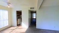 This single-floor space offers a straightforward and functional layout with distinct areas visible from a central vantage point. The living area features hard flooring bathed in natural light streaming through windows, creating a bright and open feel. A textured ceiling with a central HVAC vent provides practical climate control throughout the space.

Adjacent to the living area, the kitchen is accessible through an open doorway and is equipped with dark wood cabinetry, a white electric range with an overhead exhaust hood, and light-colored countertops. The kitchen's layout appears compact yet efficient, offering the essentials for everyday use.

A carpeted hallway extends toward the back of the space, leading to a door fitted with a window that allows additional natural light to filter in. The hallway's neutral-toned walls and carpet provide a transitional zone connecting the main living areas. Overall, the space features a neutral color palette of whites and beiges throughout, contributing to a clean and understated ambiance suited for practical, everyday living.
