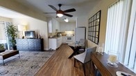 This single-floor space offers an open, flowing layout that seamlessly connects the living, dining, and kitchen areas into one cohesive environment. Rich hardwood flooring runs throughout, lending warmth and a sense of continuity to the entire space. A ceiling fan with an integrated light fixture anchors the living area, providing both airflow and ambient lighting.

The kitchen is well-appointed with white cabinetry, stone-look countertops, a tile backsplash, and white appliances including a range and refrigerator. Under-cabinet lighting adds a practical touch, illuminating the workspace effectively. The kitchen's L-shaped layout maximizes functionality while maintaining an open connection to the rest of the space.

Adjacent to the kitchen, a compact dining area features a round table with elegant X-shaped legs accompanied by upholstered gray chairs, making it an inviting spot for meals. The living area is defined by a patterned area rug and includes a dark wood dresser, adding character and storage. A large grid-style wall mirror enhances the sense of space and light. The warm, neutral wall tones throughout create a calm and welcoming atmosphere, making this a practical and comfortable single-floor home.