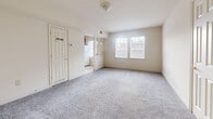 Located at 61 Shipping Pl in Dundalk, Maryland, this single-floor unit offers a practical and efficient layout comprising a living room, kitchen, and bathroom. The living room is a welcoming, open space finished with soft grey wall-to-wall carpeting and neutral beige walls. Natural light filters in through double windows, creating a bright and airy atmosphere, while a ceiling light fixture provides additional illumination. A door with glass panel inserts leads to the exterior, adding to the room's sense of openness.

The kitchen is well-appointed with warm medium-toned wood cabinetry that extends to the upper walls, providing ample storage. Light-toned countertops complement the cabinetry, and the space is equipped with a full suite of white appliances, including a gas range, dishwasher, refrigerator, and over-the-range microwave. The floor is finished with classic white square tiles, which carry through into the bathroom for a cohesive look.

The bathroom features white square floor and wall tiles, offering a clean and functional design. A rich wood-toned vanity with a countertop sink provides storage and contrast against the bright tilework, and a bathtub is visible along the wall. Overall, this unit presents a clean, functional living space with consistent finishes throughout.