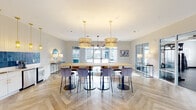 This thoughtfully designed single-story amenity facility in Kernersville, NC offers a well-rounded collection of spaces suited for socializing, working, and wellness. The central clubroom serves as the heart of the facility, showcasing elegant herringbone-patterned wood-look flooring, large statement pendant lights with gold accents, and a long communal table surrounded by plush velvet bar-height chairs. A kitchenette adjacent to this space features white cabinetry, a bold teal zellige tile backsplash, and pendant lighting with brass finishes, adding a vibrant design element. Natural light flows generously throughout via oversized glass doors and windows that frame views of the surrounding community.

A casual lounge area near the entrance features a sculptural multi-arm chandelier, soft seating with gold-legged accent chairs, and direct exterior access through full-glass double doors. A flex lounge with herringbone flooring and wainscoting provides additional open gathering space with outdoor patio access.

The fitness center is divided into two well-equipped zones: a cardio area with treadmills, ellipticals, and ceiling fans, and a strength training room featuring a dramatic galaxy-themed accent wall, free weights, benches, and cable machines. A compact co-working nook with a built-in desk, teal accent wall, and outlet access supports focused work, while a serene wellness corner provides a quiet retreat accented with colorful artwork.
