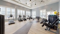 Located at 1901–1931 Hanahan Rd in North Charleston, SC, this fitness center is a thoughtfully designed, single-floor facility built to support a comprehensive workout experience. The space is divided into distinct functional zones, all unified by a clean, modern aesthetic featuring soft gray walls, light wood-style flooring, and crisp white trim.

The main workout area is generously sized and flooded with natural light from large, white-framed windows fitted with blinds. The elevated coffered ceiling with decorative shiplap panels and pendant ceiling fans adds architectural character while maintaining an open, airy atmosphere. Full-length mirrors along one wall enhance the sense of space and allow users to monitor their form. The room is equipped with a full lineup of cardio machines, including treadmills, ellipticals, and stationary bikes, as well as a dumbbell rack and adjustable weight benches.

A separate strength training zone features dedicated cable and resistance machines, stability balls, and a wall-mounted television for entertainment. This area connects to an additional room housing a dedicated cycling studio with stationary spin bikes, offering a more focused, high-intensity training environment.

Throughout the facility, light wood flooring with contrasting darker inlay strips adds visual interest, and recessed lighting ensures consistent illumination. The overall ambiance is clean, professional, and inviting, making this fitness center a well-rounded amenity for active residents.