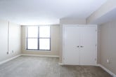 This single-floor unit offers a practical and functional layout across several interconnected spaces. The kitchen is equipped with warm-toned wooden cabinetry providing ample upper and lower storage, a tile backsplash, laminate countertops, and black appliances including a refrigerator and dishwasher. Tile flooring grounds the kitchen area, which transitions into the carpeted living space. The open connection between the kitchen and living area creates an easy flow, with natural light entering through large windows fitted with horizontal blinds. The living area is generously proportioned with neutral beige walls and overhead ventilation. The bedroom features plush carpeting, a built-in closet with shelving, and a large window allowing substantial natural light with exterior views. An additional carpeted room offers similar finishes, a full-size closet, and window access to outdoor views. The bathroom is appointed with large-format stone-look wall tiles surrounding a full soaking tub and shower combination, complemented by brushed nickel fixtures and a clean white toilet. Throughout the unit, consistent neutral tones, carpeted floors in living areas, and tile in wet zones create a cohesive and practical interior environment.
