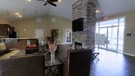 This single-floor community space makes a strong first impression with its thoughtfully designed interior that balances comfort and style. The centerpiece of the main lounge is a dramatic floor-to-ceiling stacked stone fireplace, which anchors the room and adds a rustic yet refined focal point. A flat-screen television is mounted above the fireplace, making it a natural gathering spot for residents and guests. The vaulted ceiling rises gracefully overhead, enhancing the sense of openness and volume throughout the space, while a ceiling fan provides both function and a classic finishing touch. Dark tile flooring runs throughout, grounding the room with a sleek, durable surface that complements the warm, neutral wall tones. Seating is arranged in a welcoming lounge configuration, featuring rich brown leather furniture paired with glass-top accent tables and decorative floral arrangements that add a touch of warmth. Large windows and glass entry doors along one side of the space flood the interior with natural light and provide views of the surrounding exterior. Additional rooms are accessible through interior doorways, suggesting spaces for business, recreation, or other community amenities. The overall ambiance is warm, polished, and inviting — well-suited as a central hub for community activity.