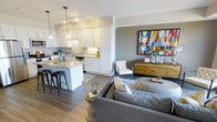 This single-floor residence showcases a seamlessly integrated open-concept layout, where the kitchen and living area flow together to create a cohesive, functional environment. The kitchen is outfitted with crisp white shaker-style cabinetry, granite countertops, and a central island with bar seating — ideal for casual dining or entertaining. Pendant lighting and under-cabinet illumination complement the track lighting overhead, ensuring the space is well-lit throughout. Stainless steel appliances and a subway tile backsplash add refined, modern touches to the culinary space. The hardwood-style flooring extends continuously from the kitchen into the living area, reinforcing the open and unified feel of the floor plan. The living space is anchored by a plush gray sectional sofa and accented with a warm wood-toned media console topped by a bold, colorful abstract artwork, introducing vibrant character against the neutral gray accent wall. A tufted accent chair adds texture and balance. The overall ambiance is contemporary and inviting, combining clean lines, warm tones, and carefully curated décor for a polished, livable aesthetic.