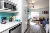 Located at 4117 University Ave in Cedar Falls, Iowa, this studio unit is a smartly designed, single-floor space that maximizes functionality within a compact footprint. The open-plan layout seamlessly connects the kitchen and living area, creating a cohesive and efficient use of space. The kitchen features white cabinetry, light countertops, a built-in microwave, stainless steel appliances including a dishwasher and full-size refrigerator, and a galley-style arrangement that keeps everything within easy reach. A bold teal accent wall energizes the main living area, where a wall-mounted flat-screen TV sits above a built-in desk with bar seating — ideal for work or dining. Track lighting runs across the textured ceiling, providing flexible illumination throughout. A standout feature is the Murphy bed, which folds away to reclaim living space when not in use, offering versatility for day-to-day living. A large window allows natural light to fill the room, complemented by a colorful area rug that adds vibrancy to the light wood-tone flooring. The bathroom is clean and functional, featuring a walk-in shower with built-in shelving and a classic navy-and-white striped shower curtain, lending a crisp, nautical feel to the space.