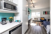 Located at 4117 University Ave in Cedar Falls, Iowa, this studio unit is a smartly designed, single-floor space that maximizes functionality within a compact footprint. The open-plan layout seamlessly connects the kitchen and living area, creating a cohesive and efficient use of space. The kitchen features white cabinetry, light countertops, a built-in microwave, stainless steel appliances including a dishwasher and full-size refrigerator, and a galley-style arrangement that keeps everything within easy reach. A bold teal accent wall energizes the main living area, where a wall-mounted flat-screen TV sits above a built-in desk with bar seating — ideal for work or dining. Track lighting runs across the textured ceiling, providing flexible illumination throughout. A standout feature is the Murphy bed, which folds away to reclaim living space when not in use, offering versatility for day-to-day living. A large window allows natural light to fill the room, complemented by a colorful area rug that adds vibrancy to the light wood-tone flooring. The bathroom is clean and functional, featuring a walk-in shower with built-in shelving and a classic navy-and-white striped shower curtain, lending a crisp, nautical feel to the space.