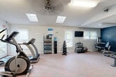 Located at 2901 Leah Court NW in Concord, NC, this fitness center offers a well-organized and comprehensively equipped exercise facility. The space features vaulted ceilings accented by dual skylights that flood the room with natural light, complemented by overhead fluorescent lighting and a ceiling fan for added comfort during workouts.

The main exercise area is finished with durable, multi-tone flooring and light-colored walls with wainscoting trim, creating a clean and functional aesthetic. A bold navy blue accent wall adds a striking visual element to one section of the room.

The facility is outfitted with a variety of cardio equipment, including multiple treadmills and an elliptical machine, along with a full cable/functional trainer machine and an adjustable weight bench. A dedicated free weights area features a dumbbell rack, kettlebells, stability balls, and a yoga mat storage rack. A wall-mounted flat-screen television and a water cooler are also provided for user convenience.

Multiple windows with blinds offer views of the surrounding outdoor greenery while maintaining privacy. The space is equipped with a keypad-secured entry door, a fire sprinkler system, and clearly marked emergency exits, ensuring safety and accessibility throughout.