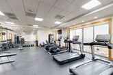 Located at 1855 Saint Francis St in Reston, VA, this fitness center offers a comprehensive workout facility within a single, open-plan space. The room is finished with durable interlocking rubber flooring in a neutral gray tone, providing a practical and comfortable surface for a wide range of exercises. A suspended drop ceiling with recessed panel lighting ensures bright, even illumination throughout the space.

Large wood-trimmed windows line the walls, allowing abundant natural light to fill the room while offering views of the surrounding area. Multiple wall-mounted flat-screen TVs are positioned throughout for entertainment during workouts.

The cardio section is well-appointed with a row of Life Fitness treadmills, elliptical cross-trainers, and a stationary spin bike, accommodating a variety of aerobic training preferences. A dedicated strength training area features a multi-station cable machine, a selectorized weight machine, an adjustable dumbbell rack with a full range of free weights, a weight bench, and various accessories including stability balls and resistance tools. A mirrored wall in the strength zone enhances the sense of space and aids in form monitoring. A beverage vending machine is also available on-site for added convenience.
