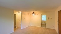 This single-floor space is characterized by its simple, functional layout and neutral aesthetic. The main living area features warm beige carpeting, white walls, and a ceiling fan with an integrated light fixture, providing both comfort and ambient illumination. A louvered closet door is visible along one wall, offering convenient built-in storage. A wall-mounted thermostat indicates individual climate control within the unit.

An open passageway leads into an adjacent carpeted room, which benefits from natural light streaming through a window with a view of surrounding greenery. This secondary space also features a baseboard heating unit, ensuring consistent warmth throughout. The open connection between the two areas creates a sense of flow and added spaciousness.

The overall design is clean and understated, with white trim, neutral carpeting, and minimal ornamentation throughout. The layout is practical and versatile, suitable for a variety of furnishing arrangements. The presence of natural light and the connection to outdoor greenery contribute to a bright and pleasant atmosphere within the space.
