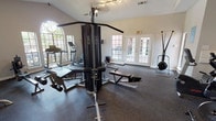 This fitness center is designed to support a comprehensive workout experience across two connected areas. The main cardio and strength room features soaring vaulted ceilings with a ceiling fan, track lighting, and an abundance of natural light streaming through large arched and multi-pane windows, as well as glass French doors that open to the outdoors. The durable rubber-speckled flooring throughout provides both safety and comfort underfoot. Cardio equipment includes a treadmill, elliptical trainer, stationary bike, and a rowing machine, while a multi-station cable machine and adjustable weight benches occupy the center of the room. A dedicated training zone is set apart by its bold orange accent wall and recessed lighting, creating an energetic and focused atmosphere. This area features a full rack of dumbbells in varying weights, kettlebells, medicine balls, a stall bar training rack, and an adjustable bench, complemented by a wall-mounted flat-screen TV for added motivation. A sanitation station is conveniently positioned near the entrance. Together, the two areas form a versatile and functional fitness facility suited for both cardio training and strength conditioning.