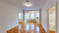 This single-floor residence showcases a cohesive layout connecting living, kitchen, and bathroom spaces in a practical and functional arrangement. The living area is the standout feature of the unit, characterized by richly toned parquet wood flooring laid in a classic block pattern that extends throughout the main rooms. A bay-style grouping of windows floods the space with natural light, while a traditional radiator beneath the windows indicates a steam heating system. Window-mounted air conditioning units provide supplemental climate control. Neutral gray-toned walls and white trim keep the interiors bright and clean throughout.

The layout, visible through the 3D floor plan, reveals an open main living zone that connects to additional rooms via wide doorways. One passage leads to a secondary room with its own window and window AC unit, along with access to a closeted area. The galley kitchen is efficiently designed with white cabinetry, dark laminate countertops, a stainless steel sink, a gas range, and a white refrigerator. Black-and-white patterned floor tile adds a classic touch. The bathroom continues this vintage aesthetic with white penny mosaic tile flooring, a soaking tub with shower, toilet, built-in shelving, and a small window for ventilation.