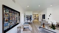 This single-floor residence showcases a thoughtfully designed open-plan layout that seamlessly connects the living, dining, and kitchen areas into one cohesive and inviting space. Light hardwood flooring runs throughout, lending warmth and continuity across every zone. The living area is anchored by plush, contemporary seating and adorned with striking large-scale artwork that gives the space a distinctly curated, gallery-like character. Recessed lighting in the clean white ceilings provides even, modern illumination that complements the natural brightness of the interior. The dining area features a round table with sleek white chairs, positioned to serve as an elegant transition point between the lounge and kitchen. The kitchen is well-appointed with stainless steel appliances, including a range and microwave, set against warm wood cabinetry and light countertops, offering both functionality and aesthetic appeal. The open sightlines from the living area through to the kitchen create a sense of spaciousness and connectivity throughout the home. Tasteful decorative accents, including sculptural pieces and framed artwork on multiple walls, contribute to an atmosphere that feels both sophisticated and livable, making this space well-suited for both everyday comfort and entertaining.