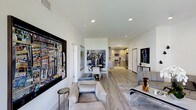 This single-floor residence showcases a thoughtfully designed open-plan layout that seamlessly connects the living, dining, and kitchen areas into one cohesive and inviting space. Light hardwood flooring runs throughout, lending warmth and continuity across every zone. The living area is anchored by plush, contemporary seating and adorned with striking large-scale artwork that gives the space a distinctly curated, gallery-like character. Recessed lighting in the clean white ceilings provides even, modern illumination that complements the natural brightness of the interior. The dining area features a round table with sleek white chairs, positioned to serve as an elegant transition point between the lounge and kitchen. The kitchen is well-appointed with stainless steel appliances, including a range and microwave, set against warm wood cabinetry and light countertops, offering both functionality and aesthetic appeal. The open sightlines from the living area through to the kitchen create a sense of spaciousness and connectivity throughout the home. Tasteful decorative accents, including sculptural pieces and framed artwork on multiple walls, contribute to an atmosphere that feels both sophisticated and livable, making this space well-suited for both everyday comfort and entertaining.
